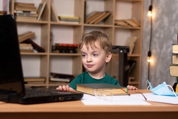 little three-year-old boy sits at the table, looks at the laptop monitor. distance learning during quarantine. next to him is a medical mask and books. the child prepares for school and does homework.