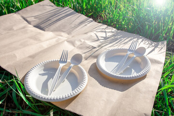 disposable biodegradable tableware on a picnic in nature. spoon, fork, plate of corn starch. green grass on the background and bright flare. modern biomaterials.