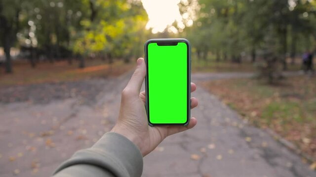Man hand holding iPhone in vertical or portrait orientation. People walking in the fall / autumn park. Pre-Keyed green screen ready for your content. POV, point of view. NY