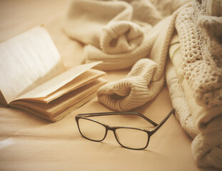 Warm sweaters are stacked against a beige background, along with an open book and black-rimmed glasses. Leisure. The comfort of home.