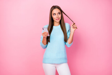 Photo of pretty blogger lady hold telephone look up empty space write new post wound curl around finger wear knitted blue sweater white trousers isolated pastel pink color background