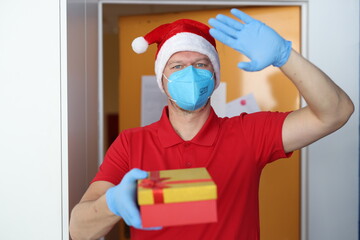 Fototapeta na wymiar Man in blue respirator and gloves hold box with gift and wave hello. Delivery of goods and gifts to door for holiday.