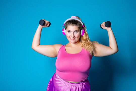 Plus size young black woman weight lifting on blue background listening music - Isolated diverse caucasian female workout with weights wearing headphones - Powered by Adobe