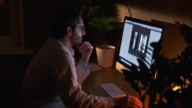 Mature and cheerful doctor with beard in shirt sitting at table looking at computer screen researching analyzes and using computer mouse in dark office room at night