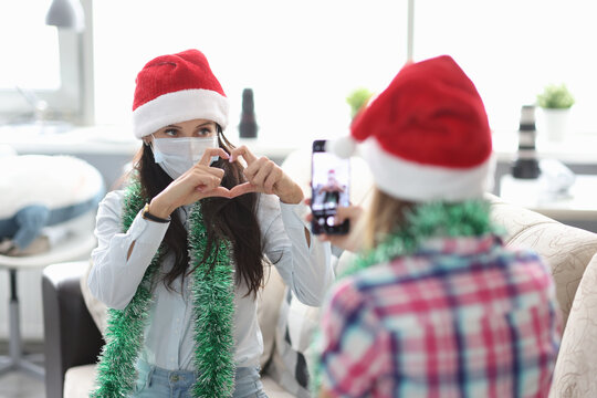 Woman in red hat and tinsel take photo on phone. Girlfriend in protective mask pose and make heart sign with hand.