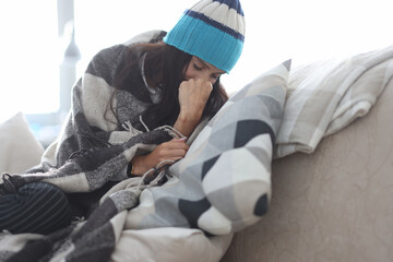 Sick woman in hat sit on sofa covered with blanket. Person on self-isolation at home.