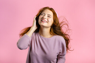 Fototapeta premium Attractive woman with red hair with headphones on pink studio background. Cute girl portrait. Music, radio, happiness, freedom, youth concept.