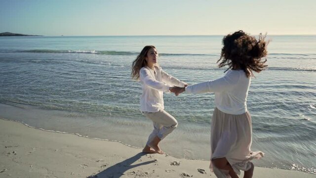 Two best female friends laughing playing ring around the rosy on the seashore with the sunset or dawn backlight - Young women having fun together running around holding hands with water reflections