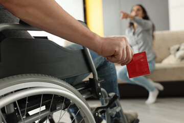 Woman sit on couch, point finger and laugh. Man in wheelchair hold red condom in his hand close up.