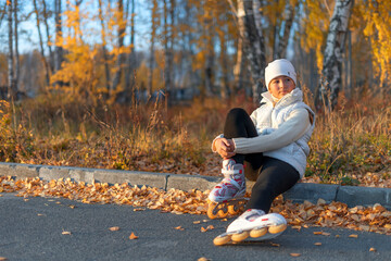A girl in roller skates is sitting on concrete curb at the park and look away. A young figure...