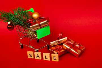 Grocery basket with golden gift boxes and Christmas balls, a sprig of Christmas tree and English letters SALE on a red background banner with copy space. Xmas sale. Christmas shopping online.