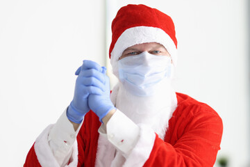 Fototapeta na wymiar Man in santa claus costume and protective medical mask and gloves stands with his hands together and smiles. Safe New Year and Christmas celebration in coronavirus pandemic concept