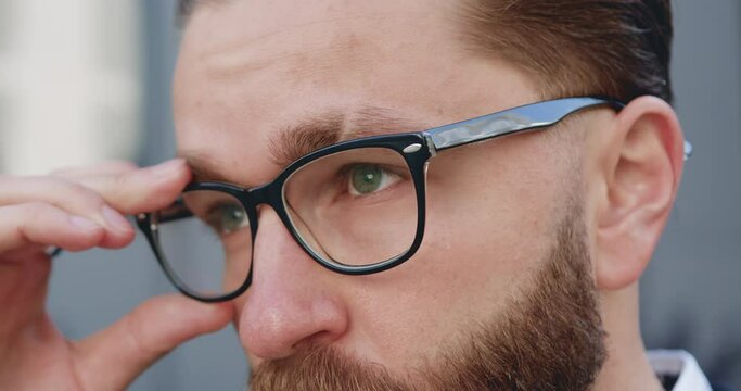 Portrait of handsome serious confident young bearded man with green eyes which putting on his glasses and looking into the distance