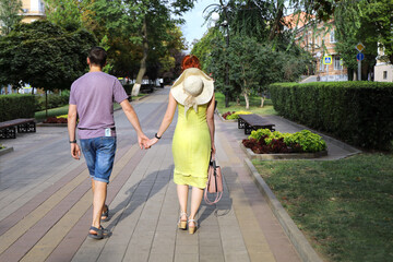 Young couple is walking holding hands along deserted alley of the city on summer sunny day. Full-length view from the back