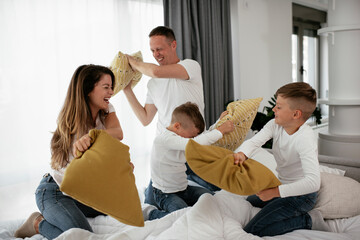 Young parents with sons fighting pillows on the bed. Happy mother and father with boys having fun at home...