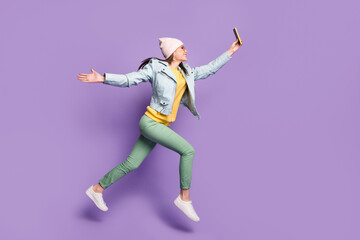 Fototapeta na wymiar Full size photo of cheerful girl jump run have free time make selfies martphone wear leather jacket green pants trousers headwear sunglass isolated violet color background