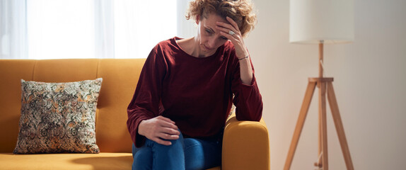 Woman with knee and headache problem sitting on a couch at home.
