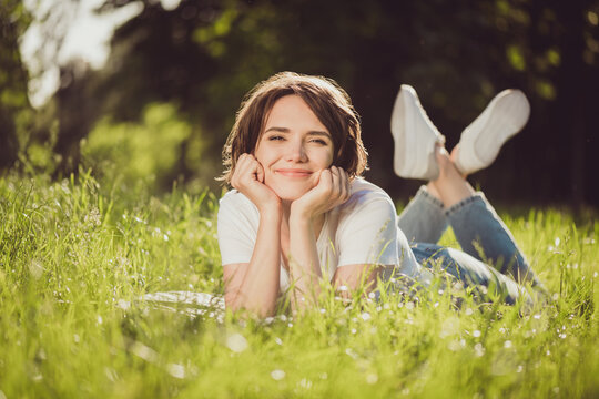 Photo of cute charming lady lying ground fresh green grass lean hands cheekbones nature harmony student resting garden overjoyed good mood wear t-shirt jeans colorful park outdoors