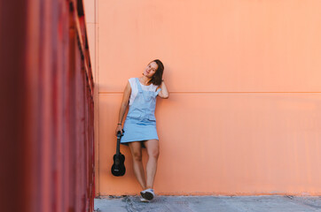 Fototapeta premium Young beautiful woman playing ukulele and having fun at the background of a coral wall
