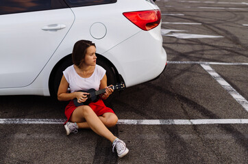 Naklejka premium Young beautiful woman sitting and having fun with ukulele near car at the parking