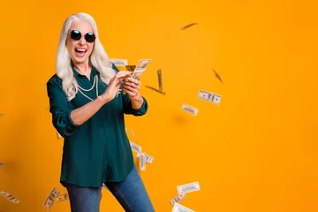 Fotobehang Photo of funky granny lady pack usa bucks hands money fall sky wealthy person spend money luxury wear green shirt sun specs necklace jeans isolated bright yellow color background © deagreez