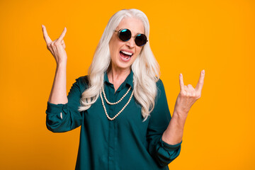 Photo of pretty crazy cheerful white haired grandma lady showing finger horns metal music lover senior party wear green shirt sun specs necklace isolated bright yellow color background