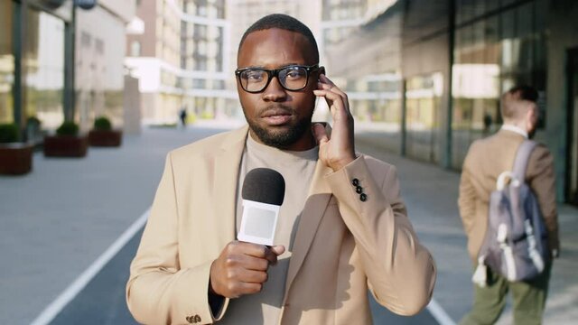 Waist up shot of Afro-American male journalist holding earphone, looking at camera and speaking in microphone while doing a live TV report outdoors in city