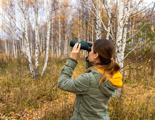 Young woman with binoculars watching birds in the autumn forest. Scientific research
