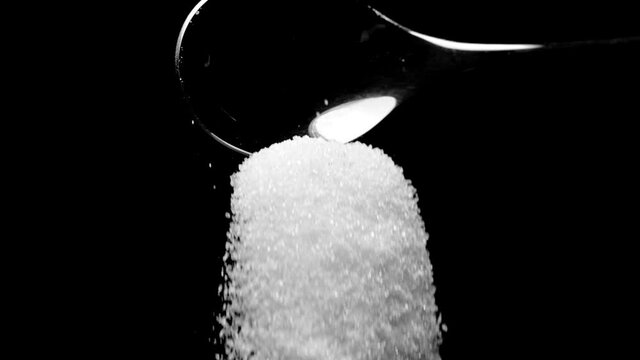White cane sugar falling down from teaspoon isolated on black background slow motion. Excess sugar leads to obesity, cardio-vascular diseases. Eat less sugar to reduce diabetes risk, healthy lifestyle