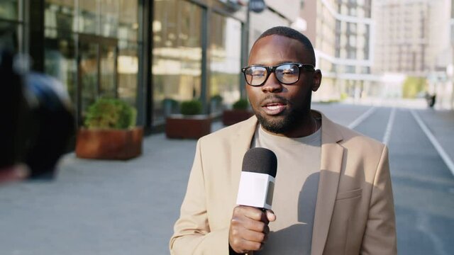 Afro-American newsman standing outdoors on urban street, holding microphone and speaking at camera while reporting in television news