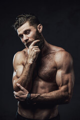 Handsome and haired man with nude torso and muscular build posing with hand under his chin in dark...