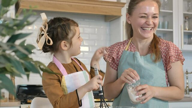 Adorable little girl with apron touches mommy nose by finger with white flour cooking cake together in contemporary kitchen