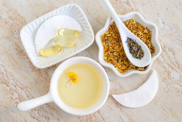 Small white bowls with cosmetic (massage, cleansing) aroma oil, dry marigold flowers, healing herbs, vitamin E capsules, eye patches. Natural skin care, homemade spa, beauty treatment recipe