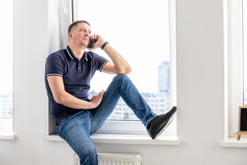 happy man talking on the phone while sitting on the windowsill of a large window
