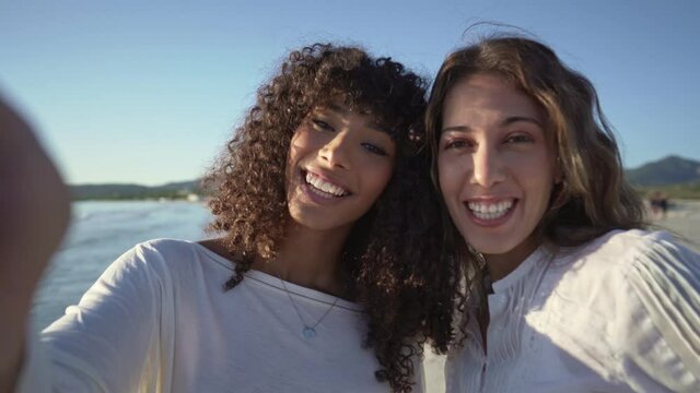 Two young cute women hugging while making a video call on seashore in vacation - Smiling multi ethnic female couple video conference from the beach at sunset or dawn