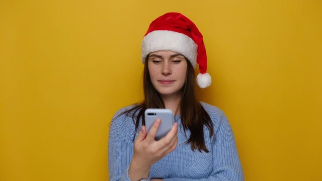 Cute young woman holds phone talks on speakerphone with friend, makes voice recognition or request uses internet services through virtual assistant, wears Christmas red hat. Translator app concept