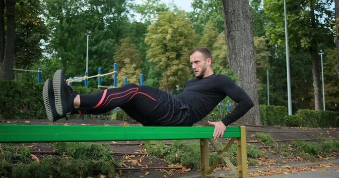 Man in sportswear doing knee tuck abdominal exercise outdoors on park bench
