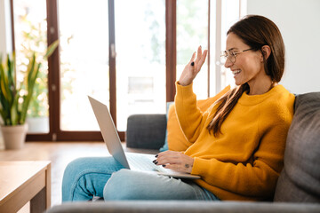 Beautiful caucasian pleased woman smiling and using laptop in apartment - obrazy, fototapety, plakaty