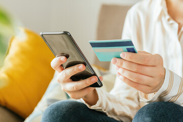 Stock photo of a young woman shopping online with her phone and a credit card from the the sofa at home