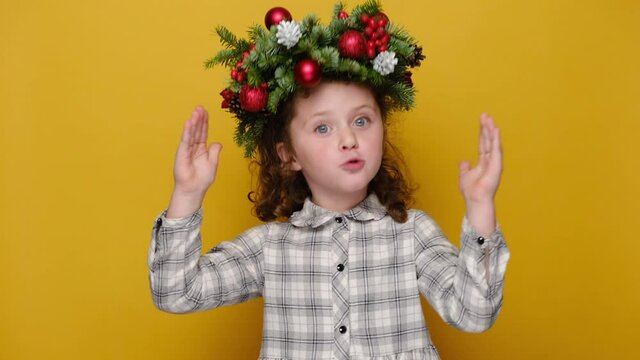 Surprised cute little kid girl showing big size gesture with hands, impressed child demonstrate something wide, wears handmade wreath, isolated on yellow studio background. Merry Christmas concept