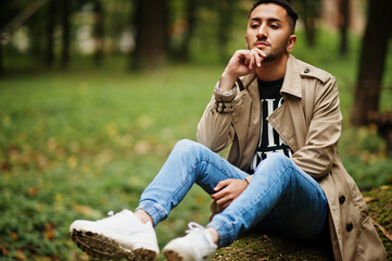 Stylish kuwaiti man at trench coat sit on tree at wood.