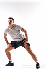 A sports man in shorts and a full-length T-shirt does exercises on a light background