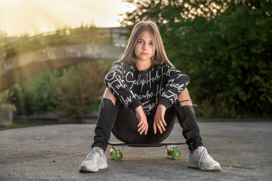 Teen girl sitting on a skateboard in the evening in the park. Stylish girl.