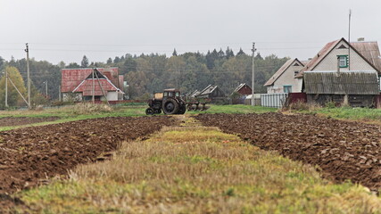 Fototapeta premium Old wheeled tractor with plow on rural brown plowed field, farming work, soil cultivation on an autumn day on forest and village houses background, beautiful countryside agriculture landscape