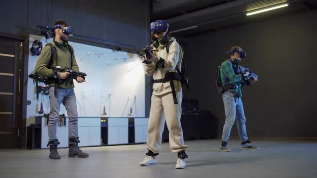 Wide full length shot of team of three young people, two men and women, in virtual reality headsets playing VR adventure game and shooting with gun controllers