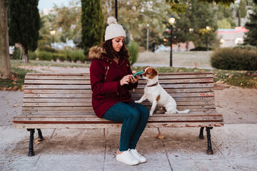 woman sitting on a bench, using mobile phone in a park with her adorable jack russell dog. Lifestyle outdoors