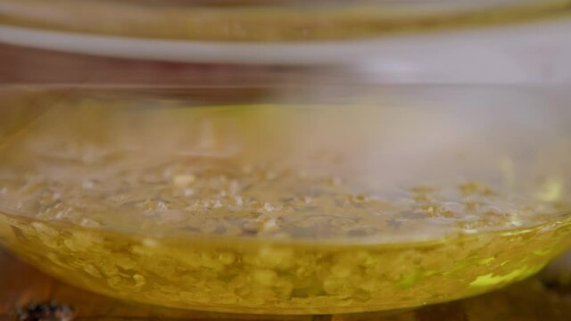 Clear transparent glass bowl on wooden table with olive oil, bottarga and fish roe dropping from top down, close up detail pan left