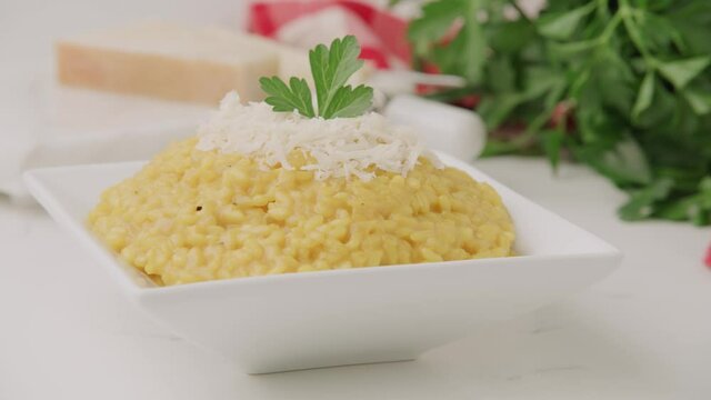Garnished, decorated, prepared, cooked yellow Italian risotto in square plate with red and white napkins, cheese and green ingredients on tablecloth table, close up pan left