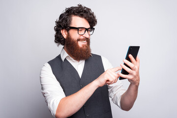 Bearded young man with glasses is smiling and typing in a tablet near a white wall