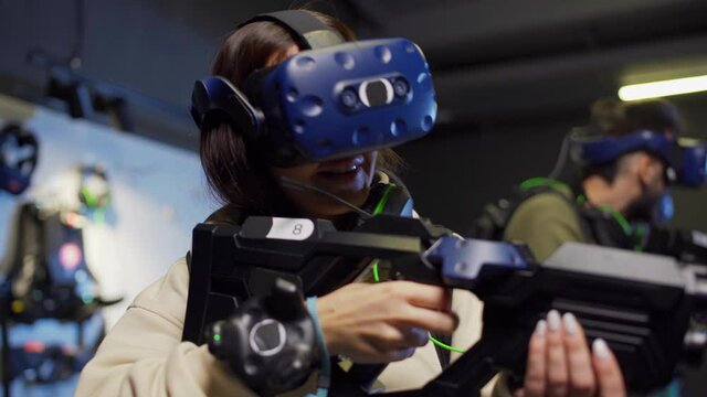 Tracking shot of group of three young people in virtual reality glasses playing VR adventure game, selective focus on young woman shooting with gun controller